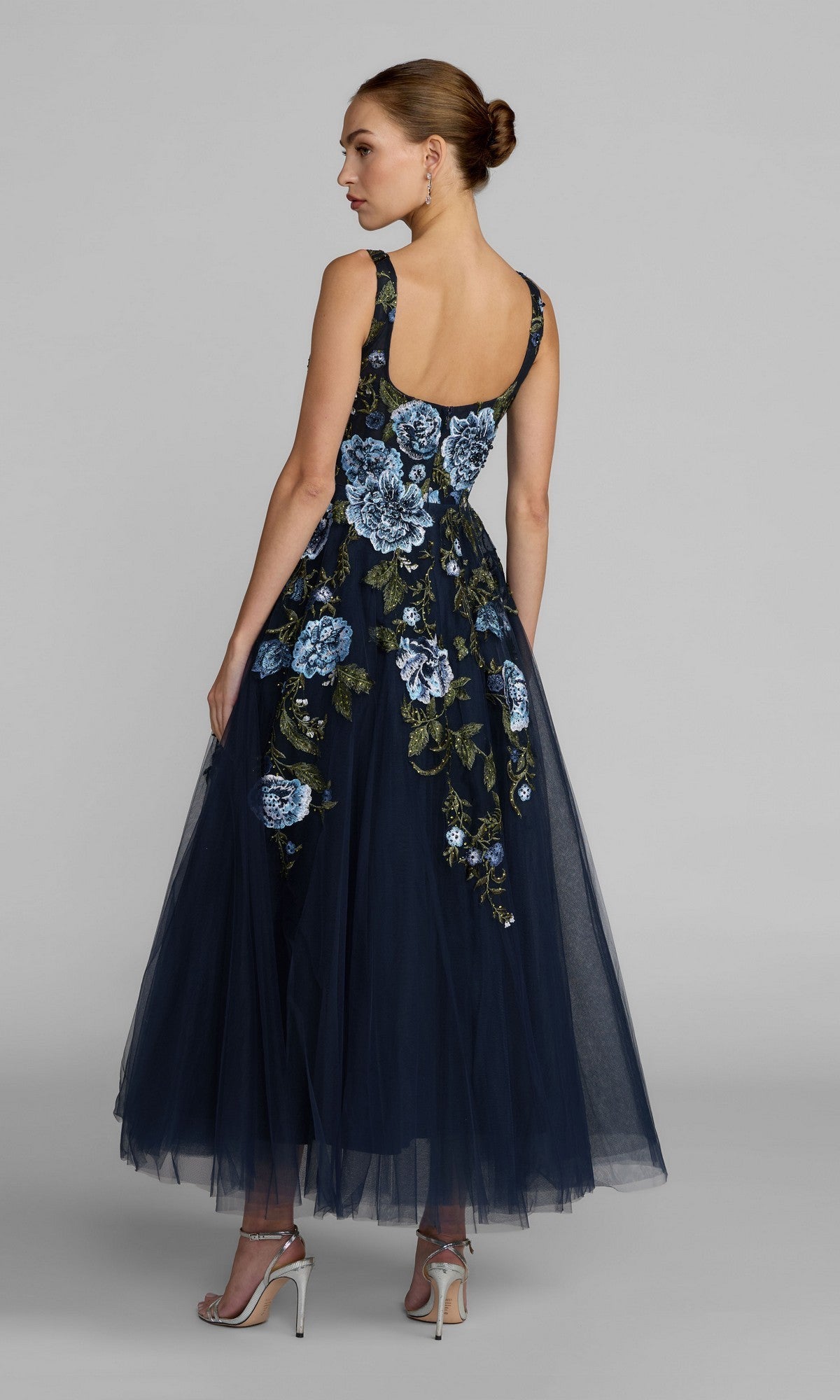 A woman stands in profile against a plain gray background, her hair styled in a bun, wearing the Tea-Length Party Dress: Mac Duggal 21295—a sleeveless navy midi dress adorned with delicate blue floral embroidery—and paired with white high-heeled sandals.